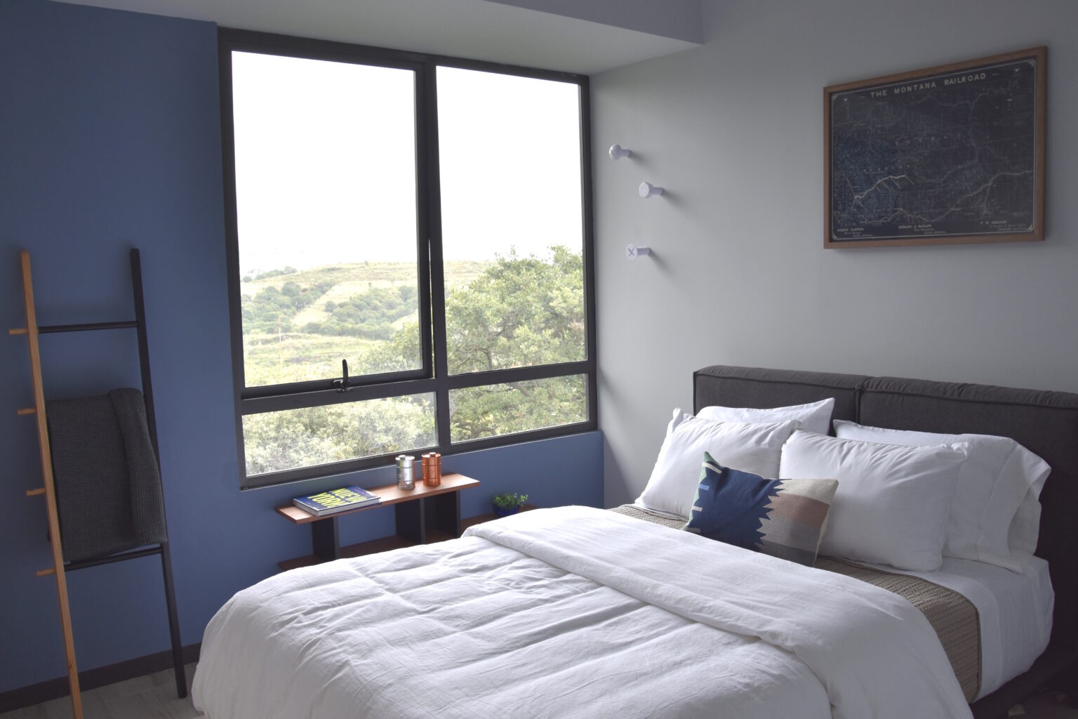 Recámara juvenil azul con cama gris, repisa de madera y vista al paisaje
