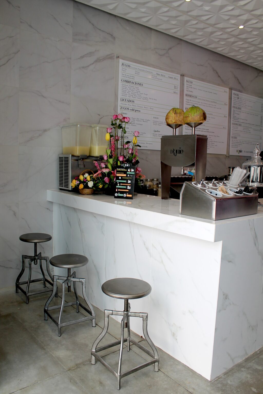 Barra blanca de mármol con bancos metálicos, menú y equipo de jugos en Dr. Juice