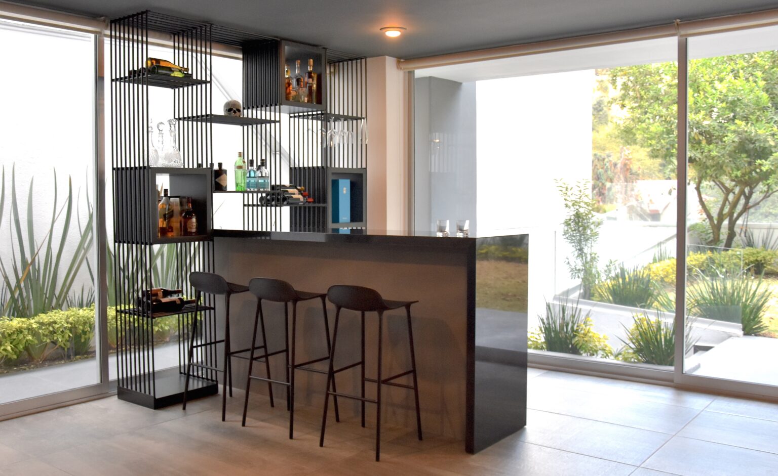 Bar residencial con barra de piedra, bancos y estantería metálica frente al jardín
