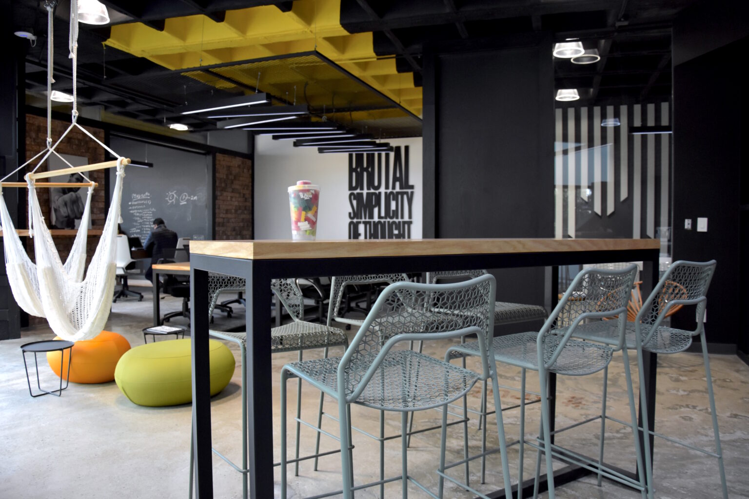 Área colaborativa con mesa alta, sillas metálicas, hamacas y plafón amarillo en M&C Saatchi
