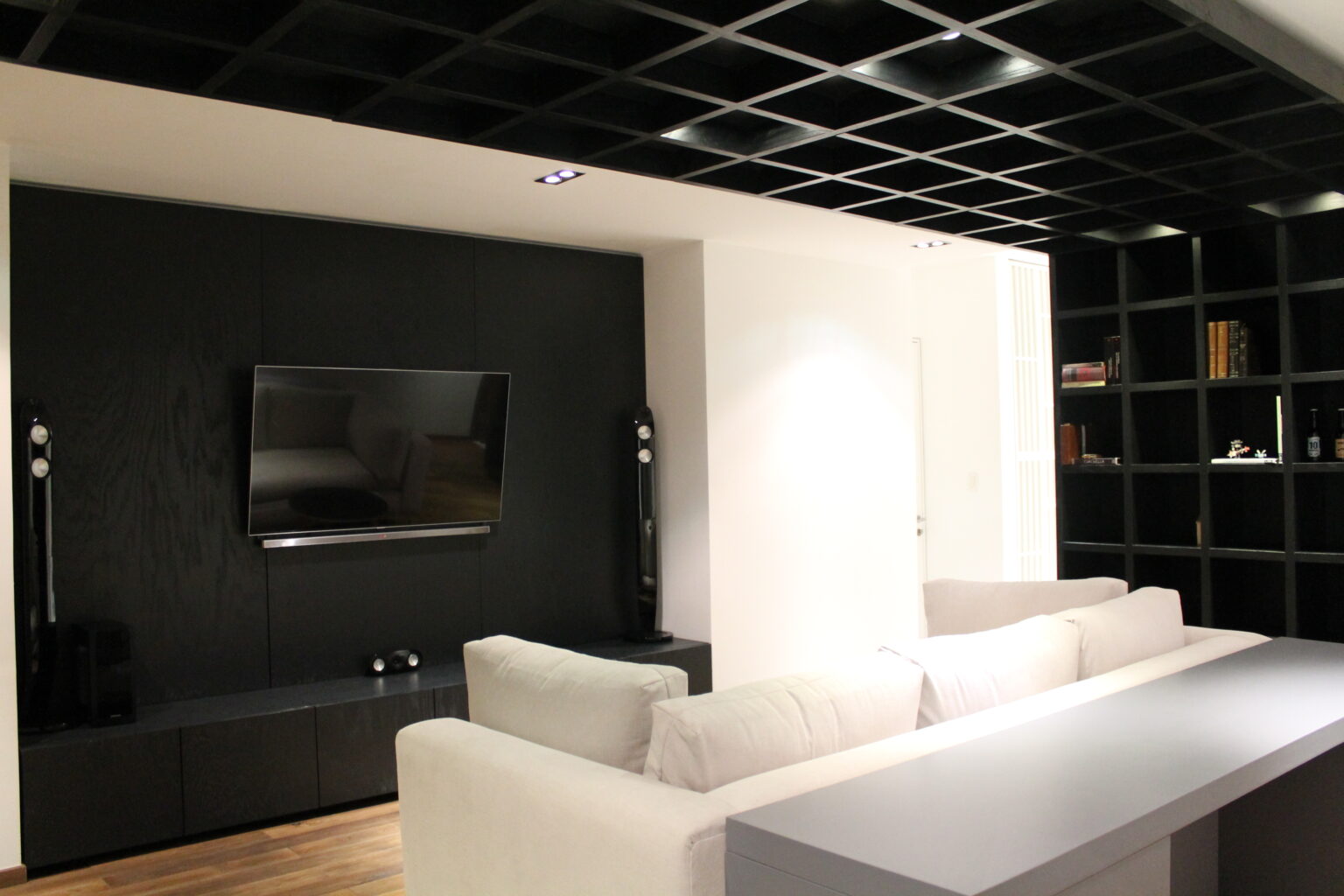 Sala de televisión de Avivia con libreros negros, plafón reticular y sofá blanco