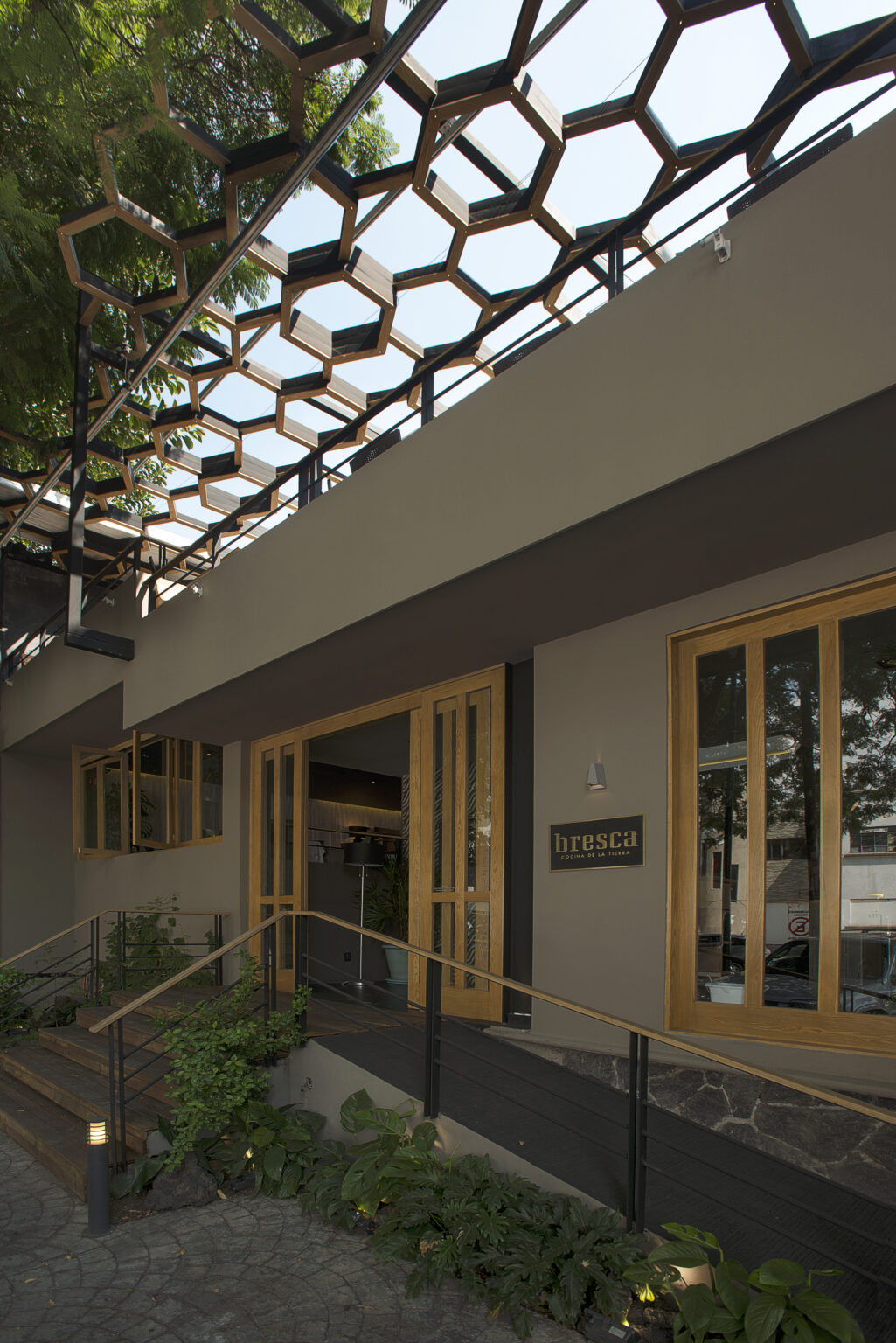 Acceso diurno a Bresca bajo pérgola geométrica con rampa y jardineras