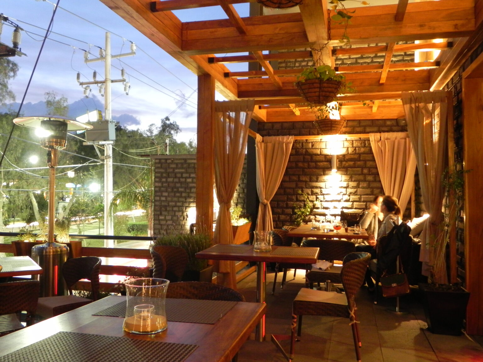 Terraza del restaurante Raíz con pérgola de madera, mesas y muro de ladrillo al anochecer