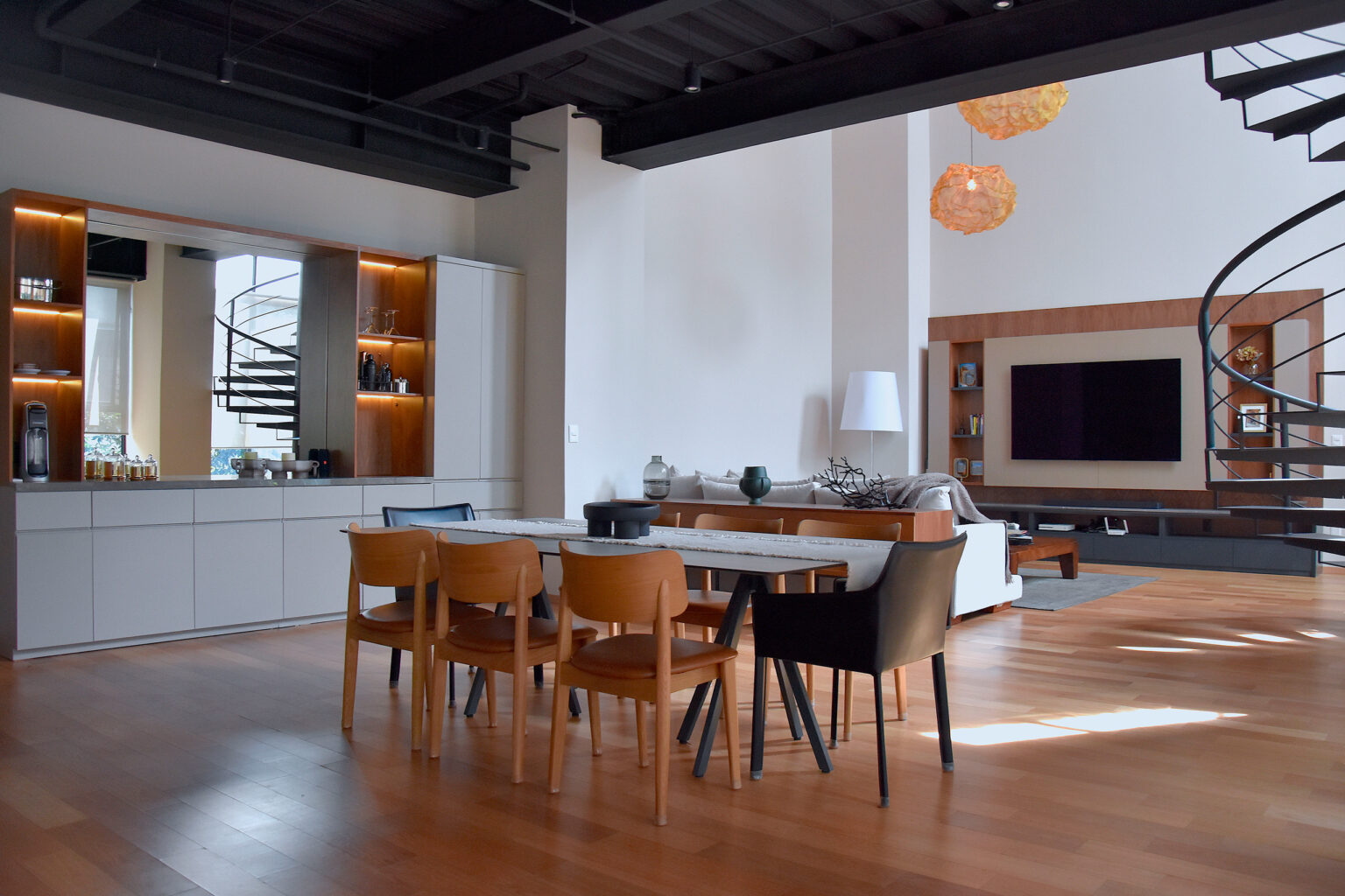 Comedor con sillas de madera frente a sala, bar iluminado y escalera helicoidal